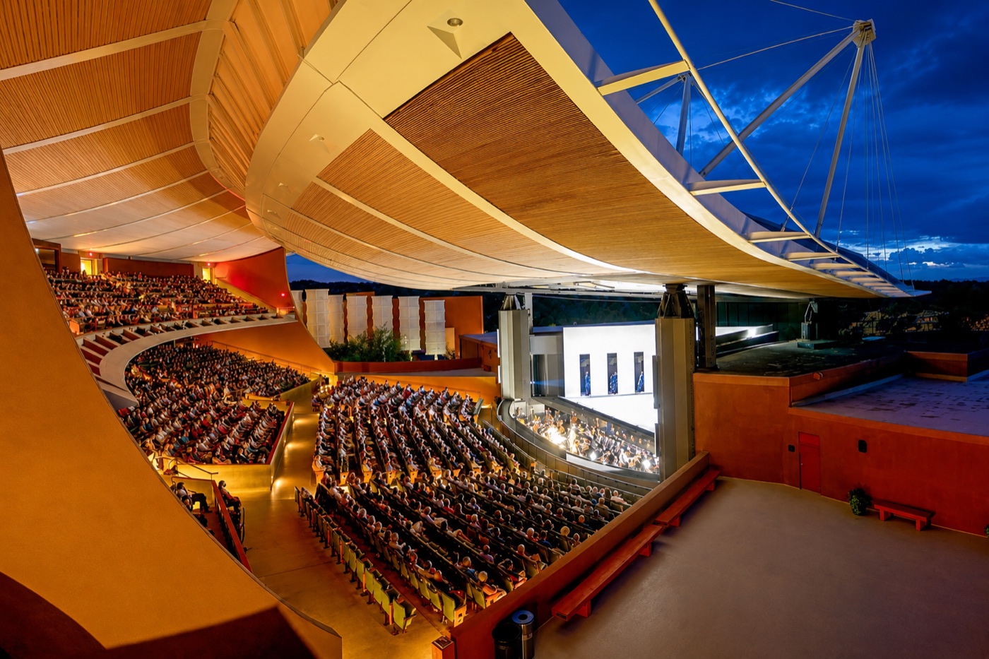 Audience filling the open-air Crosby Theatre at the Santa Fe Opera at dusk, with the curved roof canopy open to a deep blue twilight sky